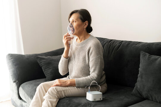 Retirement age woman using inhaler for flu and cold treatment sitting on the sofa at home, senior older female cover face with an oxygen mask. Healthcare and medicine concept