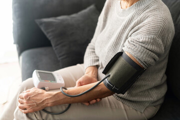 Senior woman checking blood pressure level at home, older female suffering from high blood pressure sitting at a couch and using a pulsometer, tonometer, cropped photo, face is not visible