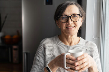 Charming elderly woman wearing stylish eyeglasses stands with cup of morning coffee looks at the...