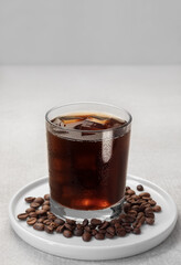 Black coffee with ice cubes in a low glass on a white saucer around coffee beans. Grey background. Water drops on a glass. Selective focus on the front wall of the glass. Copy space