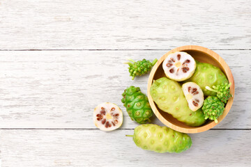 Flat lay of Noni or Morinda Citrifolia fruits in wooden bowl on white wood background.