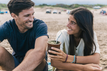 Happy young couple sharing yerba mate herbal tea at the beach.