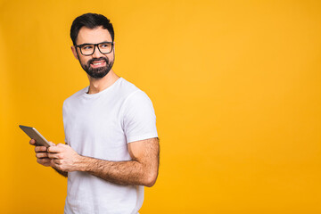 Happy young bearded man in casual standing and using tablet isolated over yellow background.