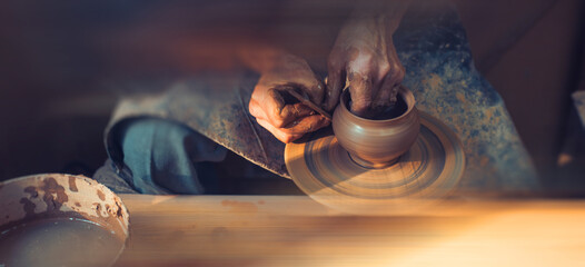 A male artisan in a pottery workshop makes a piece of clay behind a pottery wheel. Traditional pottery craft. - obrazy, fototapety, plakaty