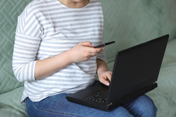 A young woman in a striped sweatshirt and blue jeans sits on the couch at home with a mobile phone in her hands and a laptop and works remotely online. Concept: freelancer. Selective focus.