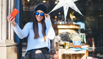 Toothy hipster girl shooting influence vlog during resting time for creating funny content, happy Caucasian blogger smiling at front camera for clicking positive selfie images via mobile app