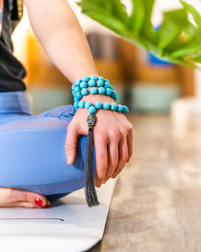 Cropped unrecognizable female in activewear sitting on mat on Half Lotus meditating with mala beads on arm in light studio