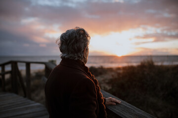 Side view of elderly female traveler in casual clothes standing on wooden pier on sandy beach and enjoying seascape at sunset