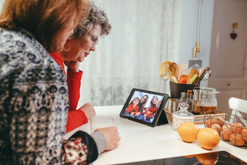 Side view of smiling homosexual elderly women watching tablet with happy children and mothers during video chat at home