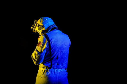 Back view of unrecognizable male cosmonaut wearing white space suit and helmet while standing on black background in blue and yellow neon light