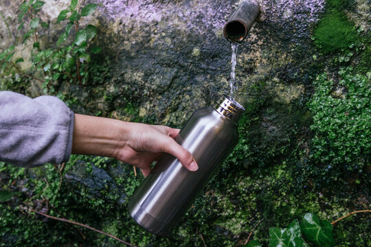 Crop anonymous hiker filling metal bottle with fresh cold natural spring water from source with pipe in mountainous forest