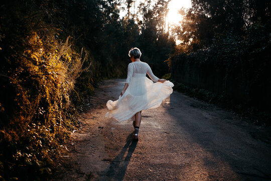 Full Body Back View Of Anonymous Female Wearing White Dress Walking On Rural Road Among Green Trees In Nature On Evening Time