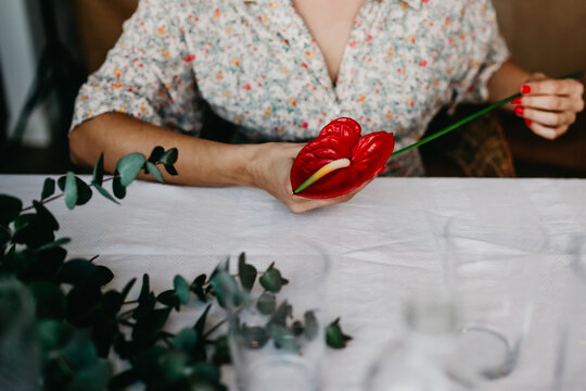 Unrecognizable female with red flower pestle sitting at table with white tablecloth near green foliage while cultivating plant at home