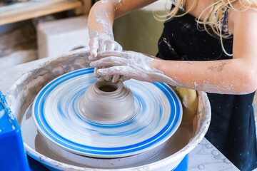 Crop unrecognizable little girl in black apron standing near pottery wheel while shaping clay pot in light workshop