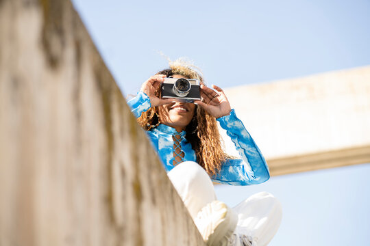 From below of happy curly haired ethnic female in stylish outfit sitting on concrete fence and taking pictures with retro photo camera in sunny summer day