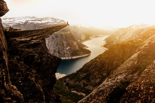 Distance view of explorer sitting on edge of Trolltunga rock formation at sunset during vacation in Norway