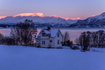 Lonely residential cabin located on shore of lake on background of snowy mountain range lit by pink sunset light in winter in Norway