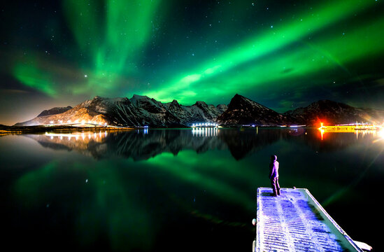 Unrecognizable person standing on pier and admiring scenery of green polar lights glowing in night starry sky above mountains in winter in Norway