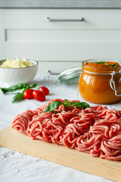 From above of fresh raw mixed pork and beef minced meat on wooden cutting board with green basil leaves placed on table during cooking process near homemade tomato sauce