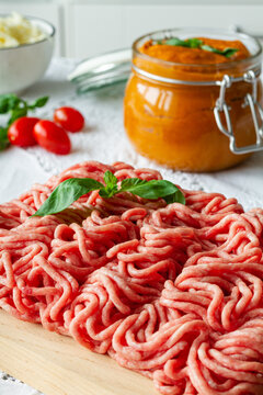 From above of fresh raw mixed pork and beef minced meat on wooden cutting board with green basil leaves placed on table during cooking process near homemade tomato sauce