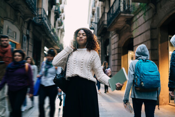 Beautiful dark skinned female in trendy wear standing on crowded street concentrated on favorite songs in earphones, pensive dark skinned woman in headphones enjoying online music outdoors