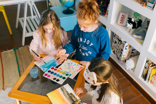 Cute adorable girls sitting at table with sheets of paper and watercolor while drawing pictures together