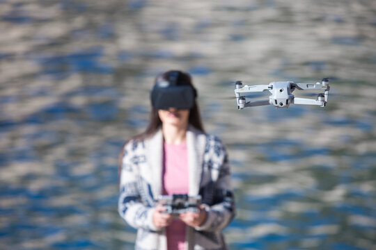 Excited female wearing modern VR goggles operating drone with remote controller and experiencing virtual reality while standing against blurred sea