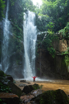 Back view of unrecognizable traveler in warm clothes standing in pond water with outstretched arms near picturesque Haew Narok Waterfall streaming through rocky cliff covered with lush green tropical vegetation