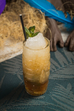 From above of tiki cup with cold alcohol drink with straw served with ice and decorated with fresh herb placed against dry grass on blurred background