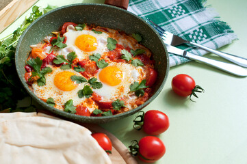 Traditional dish of israeli cuisine Shakshuka. Fried eggs with tomatoes, peppers, onions, cilantro and pita in a pan on light green background