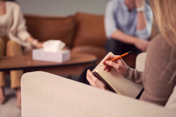Unrecognizable female psychologist talking notes in notebook during therapy session with couple in office