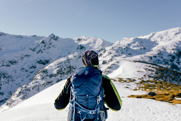 Back view of unrecognizable male mountaineer in warm activewear with backpack standing on slope of snowy rocky mountain and enjoying spectacular landscape in sunny winter day