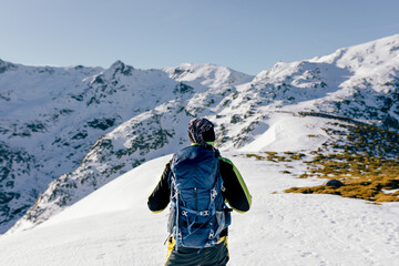 Back view of unrecognizable male mountaineer in warm activewear with backpack standing on slope of snowy rocky mountain and enjoying spectacular landscape in sunny winter day