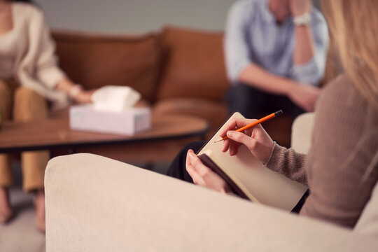 Unrecognizable female psychologist talking notes in notebook during therapy session with couple in office