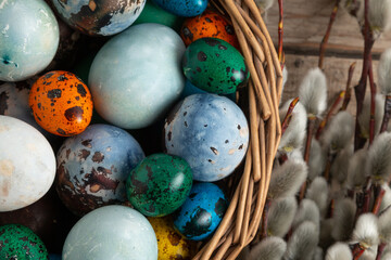 Various colorful and beautiful Easter eggs are placed in a wicker basket on a wooden table. Willow branches in the background