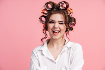 Portrait of positive girl winking isolated over pink background. Cute, cheerful girl with curlers...