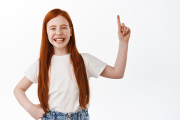 Cheerful little red head girl, kid with ginger hair pointing finger up and laughing over funny...