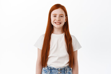 Cute little kid with long red hair smiling and looking happy at camera, standing over white background. Girl child express joy and positive emotions - 423082558