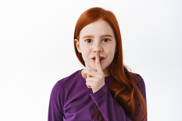 Shh this is secret. Cute redhead little girl, child shushing, show keep quiet taboo gesture, finger on lips, hushing, standing over white background