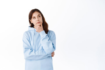 Portrait of brunette girl with thoughtful face, touching chin and looking aside at upper right corner, making choice, shopping and thinking, standing against white background