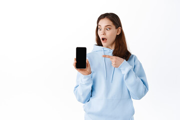 Naklejka premium Shocked young woman pointing finger at her phone screen, gasping amazed while staring at smartphone display, standing against white background