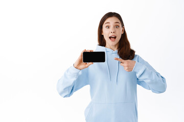 Excited girl pointing and showing smartphone screen flipped horizontally, demonstrates an application, standing against white background