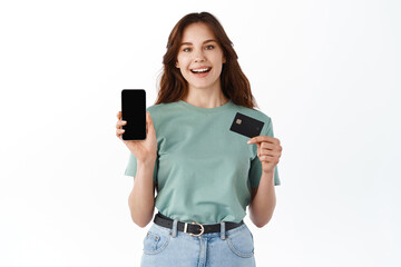Fototapeta premium Portrait of a happy young brunette girl showing plastic credit card while holding mobile phone isolated over white background