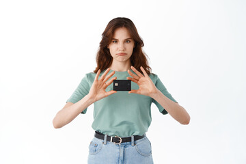 Sad young woman in t-shirt showing her plastic credit card and grimacing upset, having bad mood,...