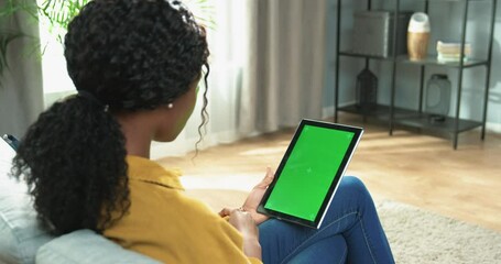 Over shoulder view of African American woman sitting on sofa in apartment in living room tapping on tablet device with green screen with tracking motion browsing online, technology concept - Powered by Adobe