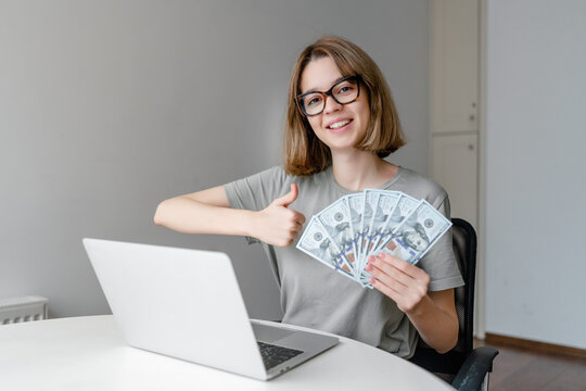 young caucasian woman showing money and thumb up sitting with laptop at home
