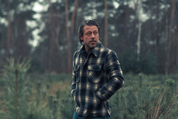 Man with stubble beard in a checkered coat in a rainy woodland.