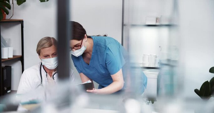 Medics work together. Nurse woman in blue scrubs gives papers to senior Caucasian grey haired doctor woman in lab coat.