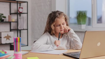 Cute primary school girl studying at home using laptop computer. Schoolgirl has online lesson, writing in exercise book, doing homework. Distance learning online. Children home education