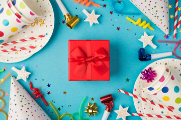 Top view photo of red gift box with ribbon bow in the middle of birthday table composition and party accessories on isolated blue background